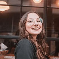 Uma mulher, com idade entre 20 e 25 anos, está sorrindo enquanto olha para a foto. Ela usa uma blusa preta, tem cabelo longo e está de óculos